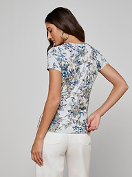 A woman with long brown hair wears large hoop earrings and the Ressi Fitted Tee with a butterfly print, paired with white pants, standing with her back to the camera against a plain light gray background.