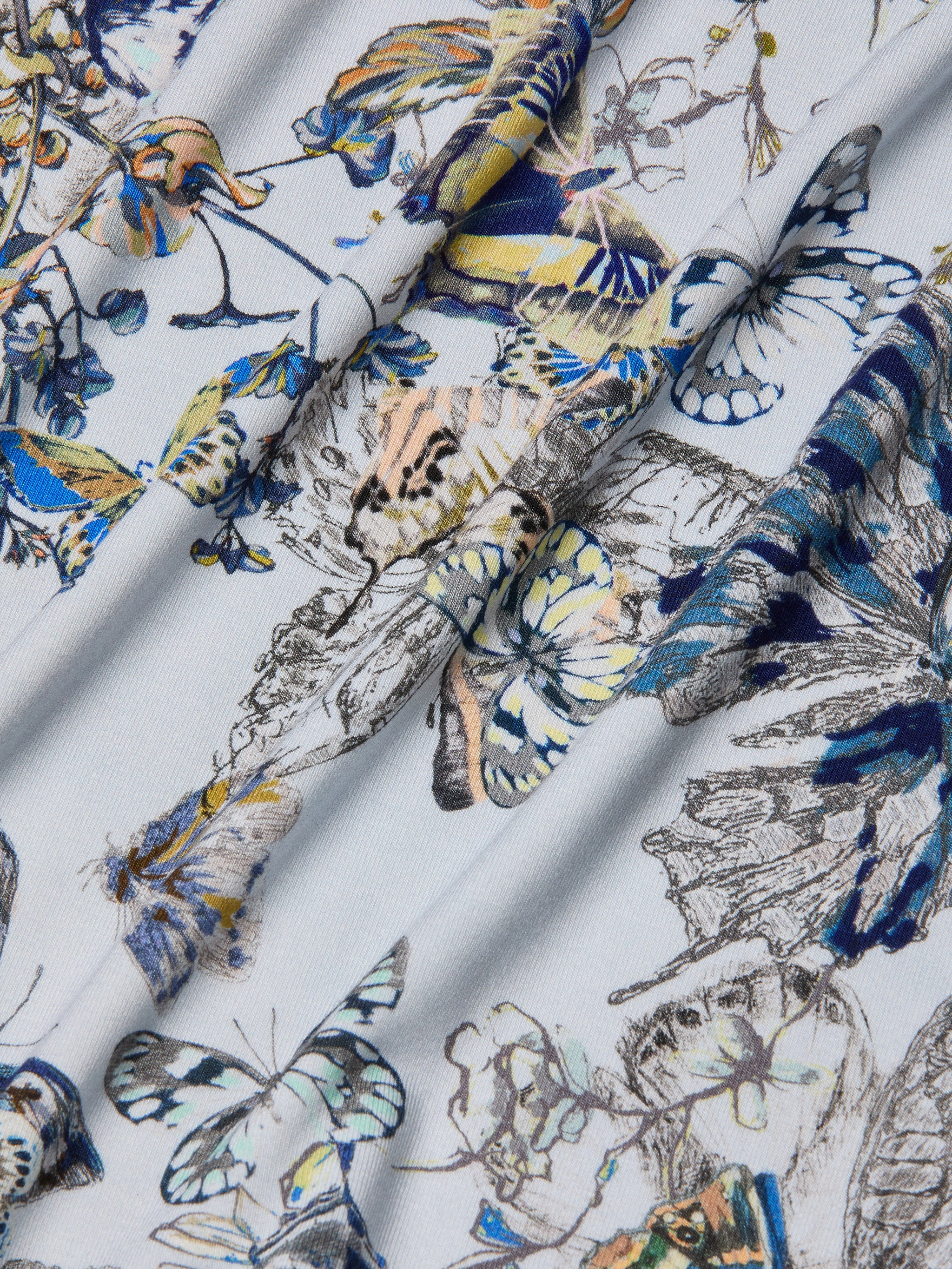 A close-up of stretch jersey fabric with a colorful butterfly print in blue, yellow, and beige on a light background. The softly folded fabric highlights the texture and pattern, perfect for the Ressi Fitted Tee.