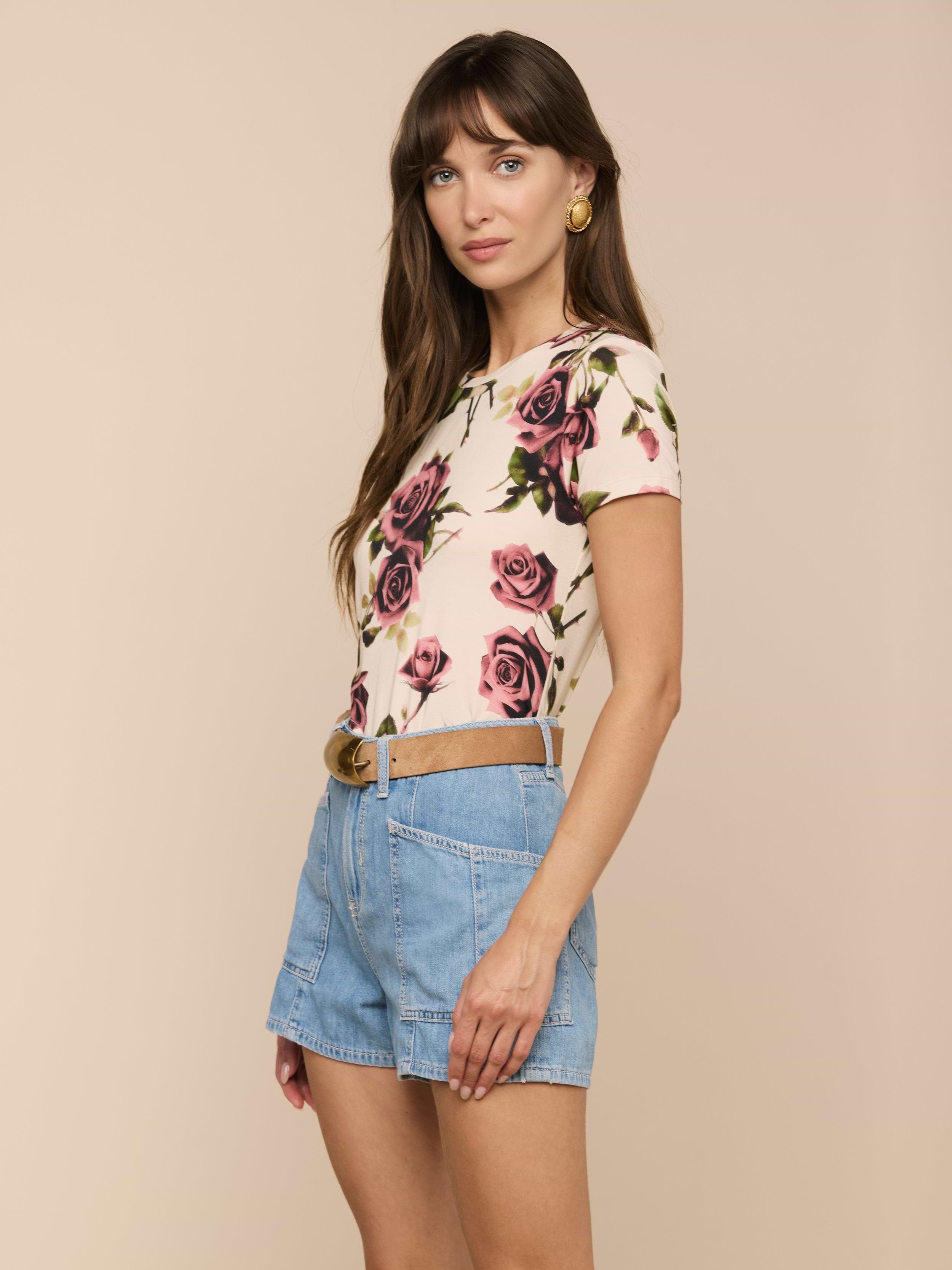 A woman stands sideways against a plain background, looking at the camera, wearing the Ressi Fitted Tee—a light-colored floral top with pink roses crafted from stretch jersey knit—paired with denim shorts and a belt.