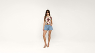 Woman wearing sunglasses, a floral-print shirt, blue shorts, and high-heeled sandals stands against a plain white background with one hand in her pocket.