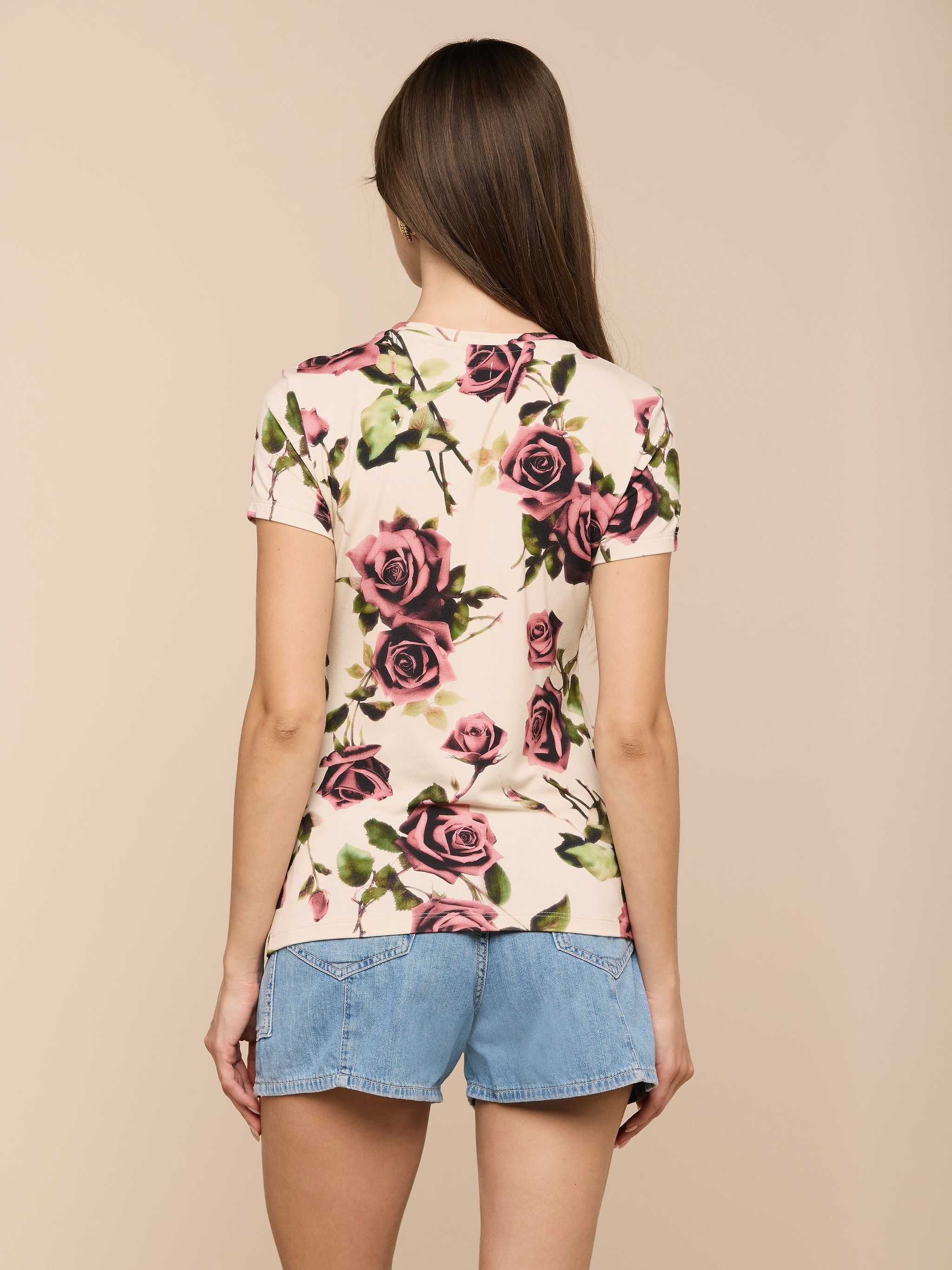 A person with long brown hair wears the Ressi Fitted Tee, a slim-cut, rose-patterned short-sleeve T-shirt made from stretch jersey knit, paired with denim shorts, standing with their back to the camera against a light background.