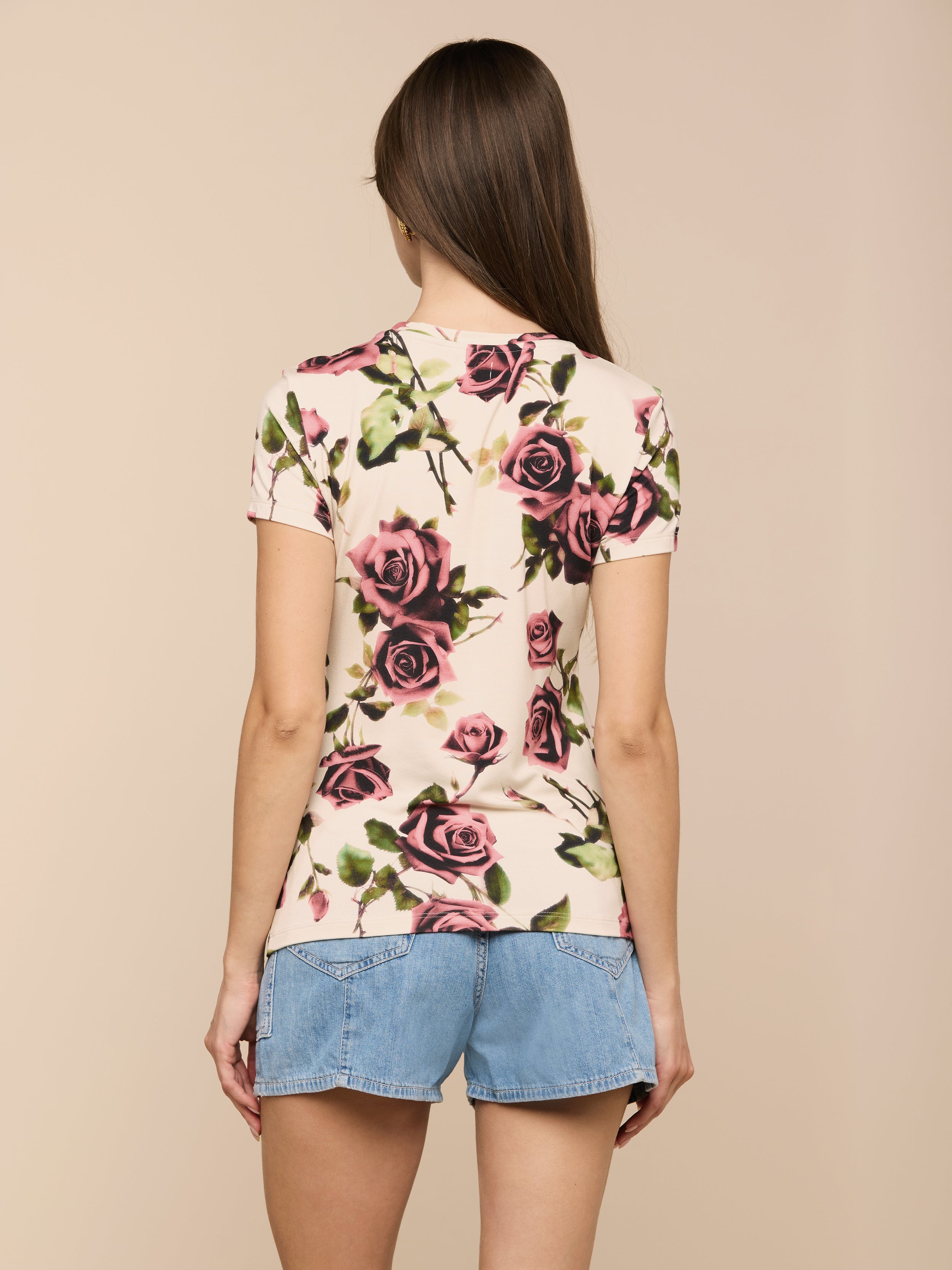 A person with long brown hair wears the Ressi Fitted Tee, a slim-cut, rose-patterned short-sleeve T-shirt made from stretch jersey knit, paired with denim shorts, standing with their back to the camera against a light background.