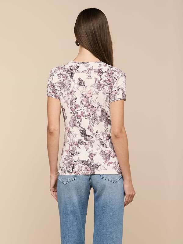 A woman with long brown hair wears the bestselling Ressi Fitted Tee in a light butterfly print and blue jeans, standing with her back to the camera against a beige background.