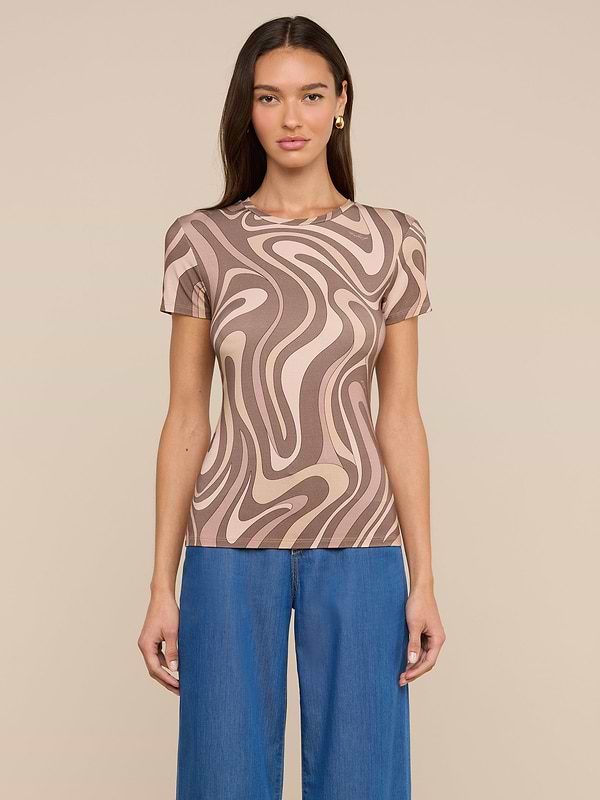 A woman stands against a light background wearing the Ressi Fitted Tee—a short-sleeve, beige and brown abstract swirl print top in stretch jersey knit—paired with blue jeans. She has long, straight brown hair and faces forward with a neutral expression.