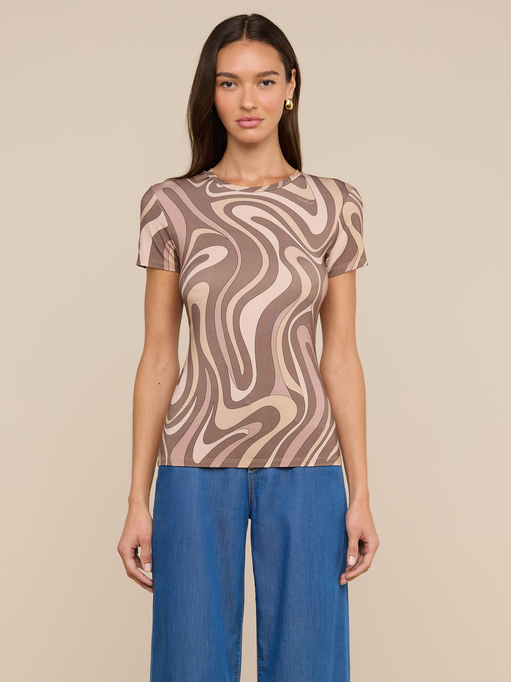 A woman stands against a light background wearing the Ressi Fitted Tee—a short-sleeve, beige and brown abstract swirl print top in stretch jersey knit—paired with blue jeans. She has long, straight brown hair and faces forward with a neutral expression.