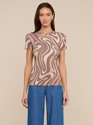 Une femme aux longs cheveux bruns porte le Ressi Fitted Tee, un best-seller en jersey stretch à la coupe ajustée, avec un motif ondulé beige et marron, associé à un jean bleu. Elle se tient de face sur un fond beige uni.