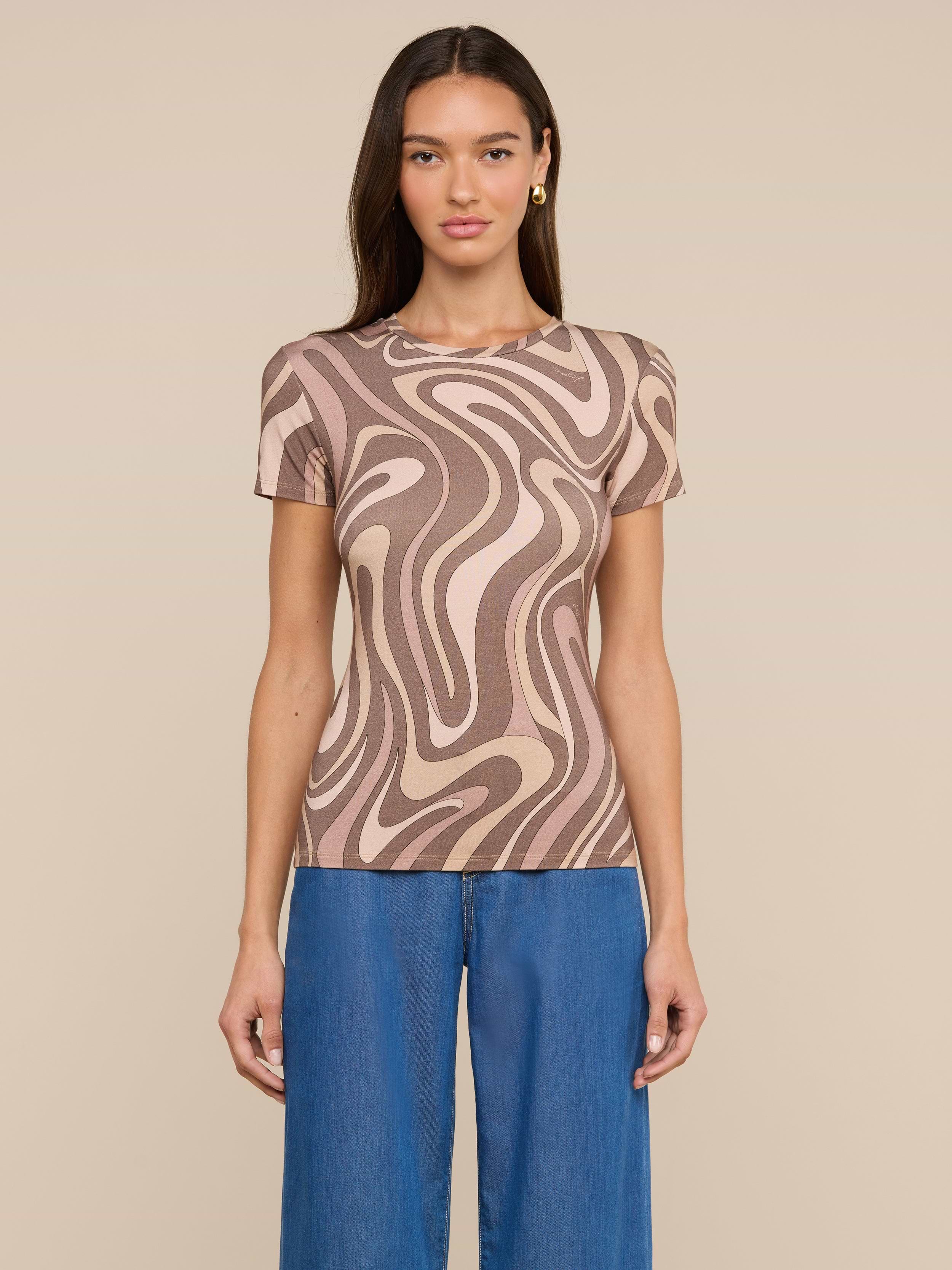 A woman stands against a light background wearing the Ressi Fitted Tee—a short-sleeve, beige and brown abstract swirl print top in stretch jersey knit—paired with blue jeans. She has long, straight brown hair and faces forward with a neutral expression.