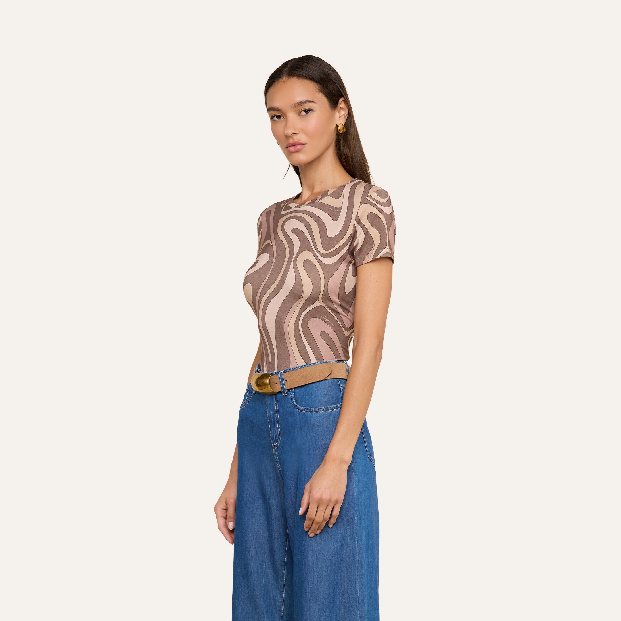 Woman wearing the Ressi Fitted Tee, a short-sleeve stretch jersey knit top with an abstract swirl print in brown and beige, paired with blue wide-leg jeans and a brown belt, standing against a plain off-white background.