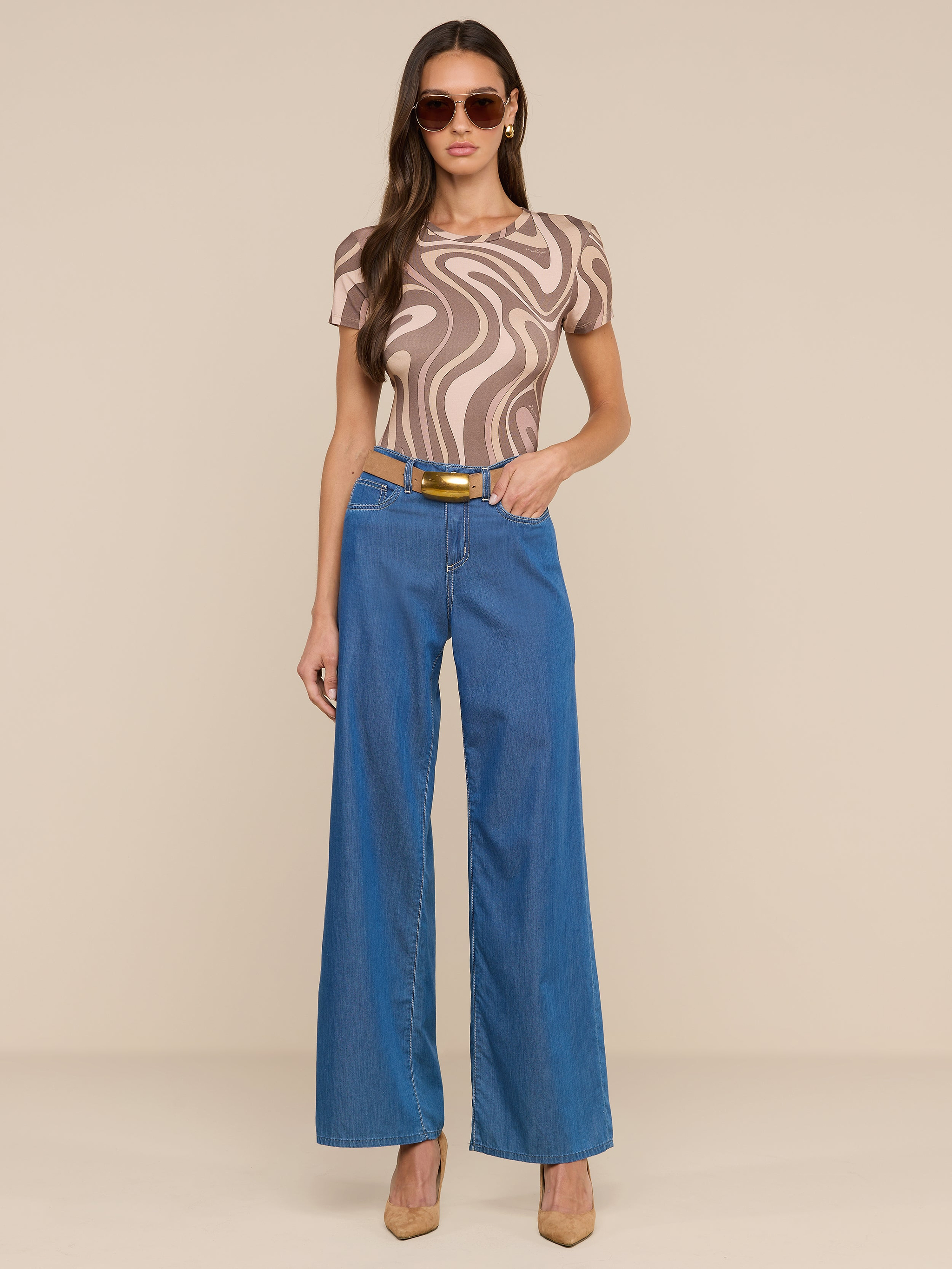A woman wearing sunglasses, the Ressi Fitted Tee—an abstract swirl print beige and brown patterned top—wide-leg blue jeans with a belt, and tan heels stands against a plain white background.