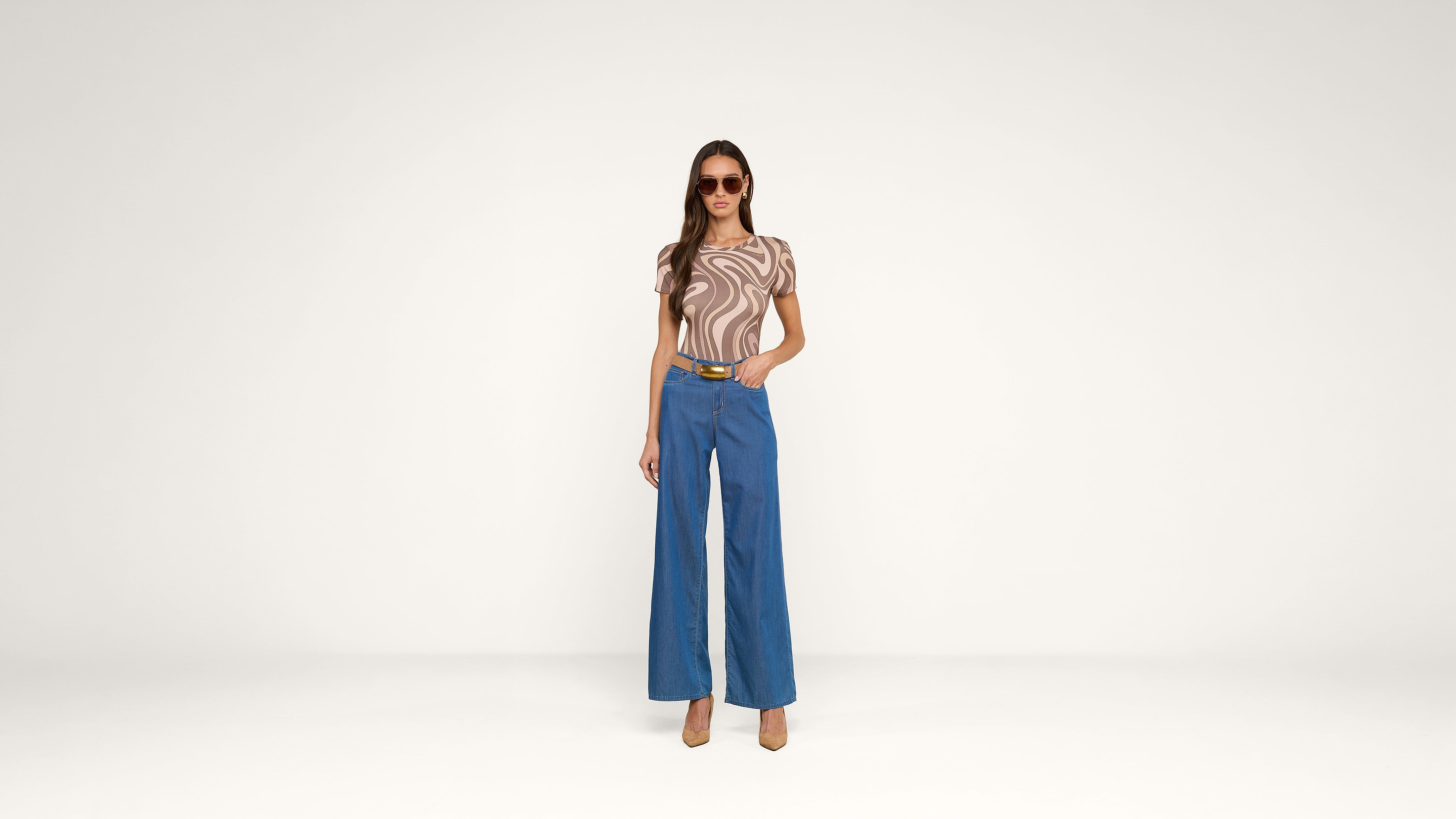 A woman wearing sunglasses, a brown and beige swirled top, blue wide-leg pants, and heels stands confidently against a plain white background.
