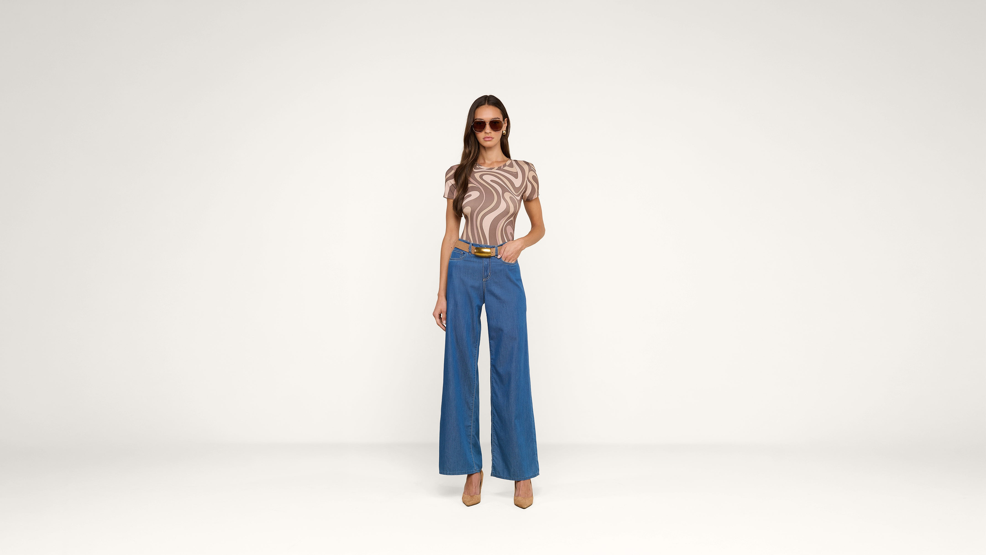 A woman wearing sunglasses, a brown and beige swirled top, blue wide-leg pants, and heels stands confidently against a plain white background.