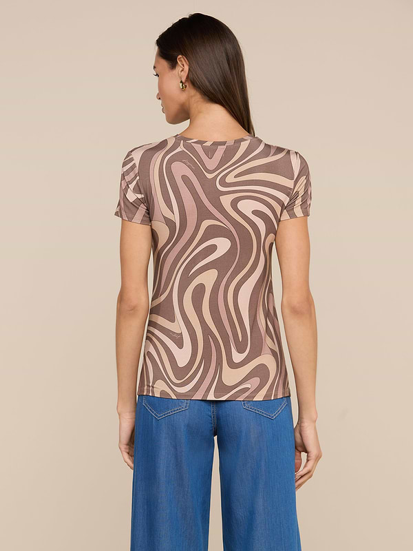 A woman stands facing away, wearing the bestselling Ressi Fitted Tee featuring an abstract swirl print in brown and beige. She pairs it with wide-leg blue jeans. Her straight hair is down, and she has small hoop earrings. The background is plain and light-colored.