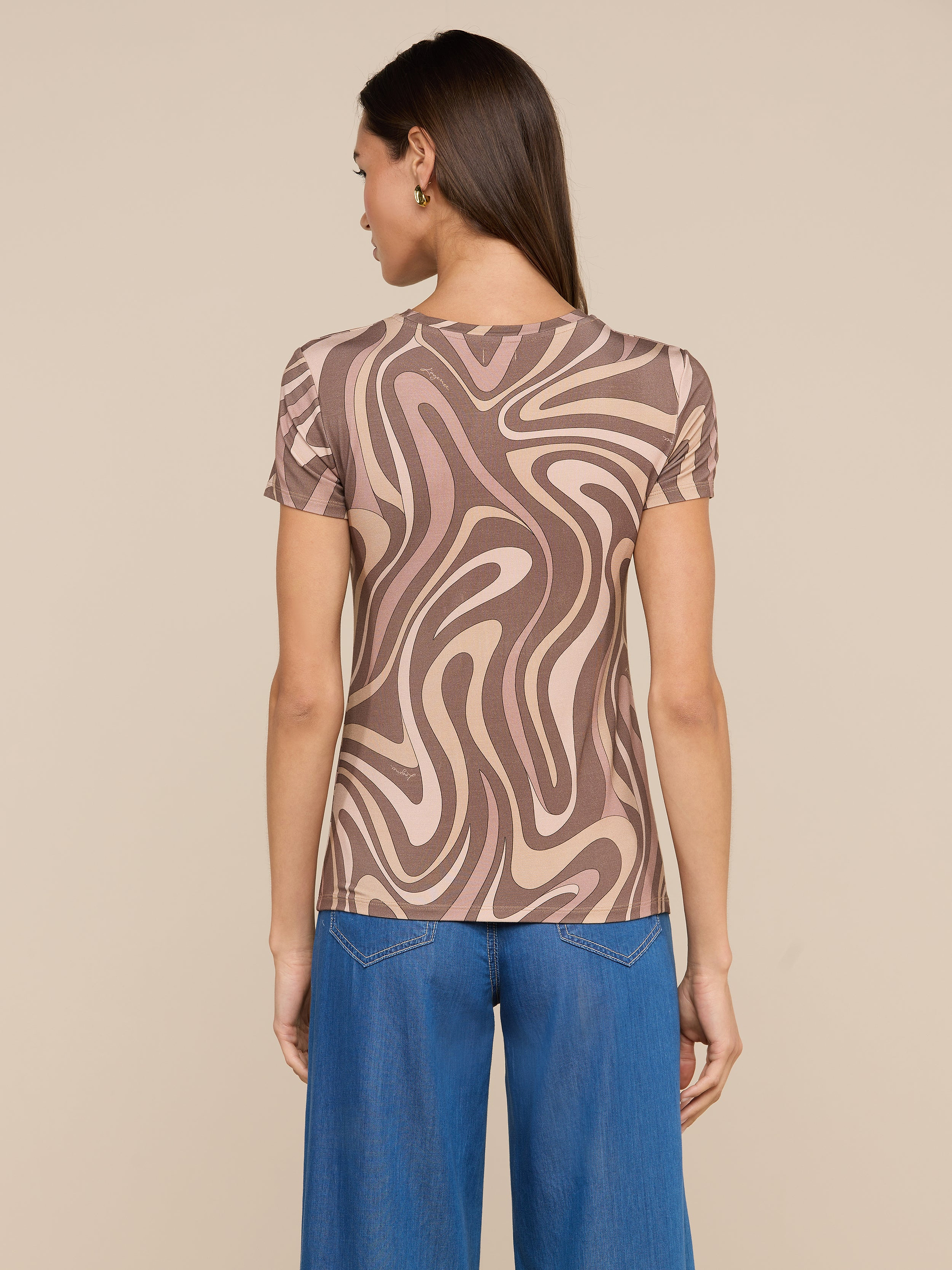 A woman stands facing away, wearing the bestselling Ressi Fitted Tee featuring an abstract swirl print in brown and beige. She pairs it with wide-leg blue jeans. Her straight hair is down, and she has small hoop earrings. The background is plain and light-colored.