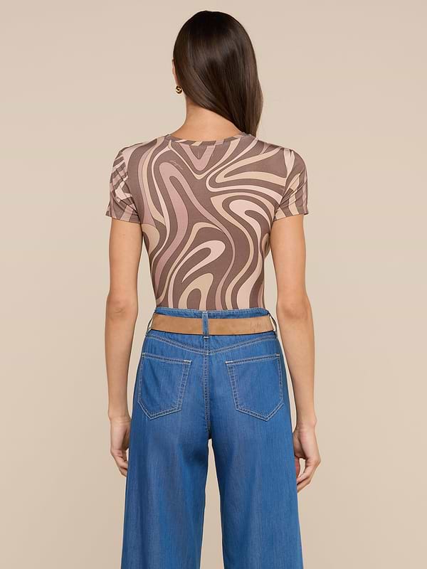 A woman faces away, wearing the bestselling Ressi Fitted Tee featuring an abstract swirl print in brown and beige, paired with high-waisted wide-leg blue jeans. Her straight brown hair falls over her shoulder against a simple off-white background.