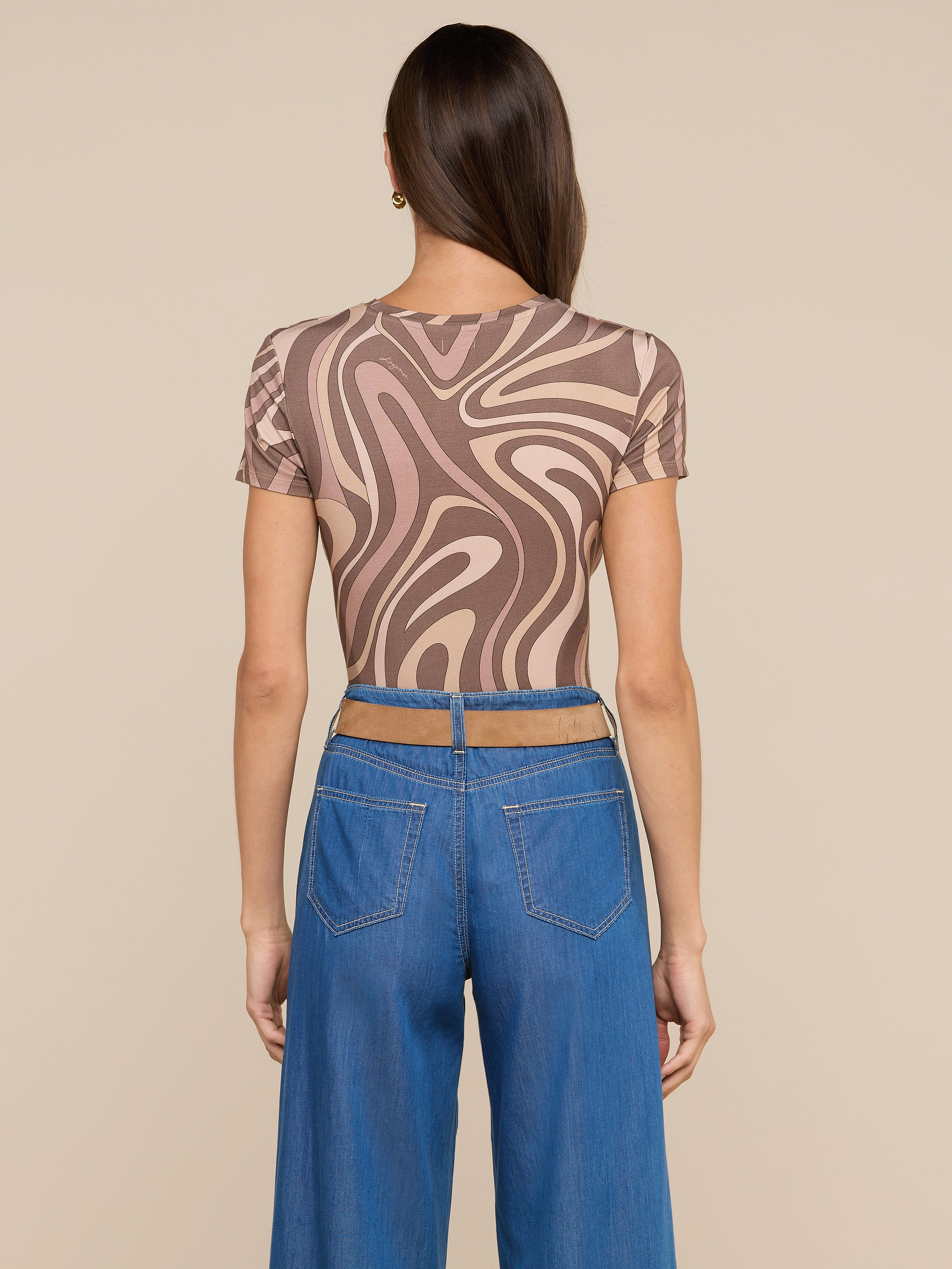 A woman faces away, wearing the bestselling Ressi Fitted Tee featuring an abstract swirl print in brown and beige, paired with high-waisted wide-leg blue jeans. Her straight brown hair falls over her shoulder against a simple off-white background.