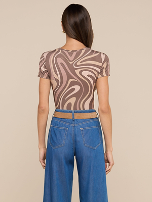 Une femme aux longs cheveux bruns est vue de dos, portant le tee-shirt ajusté Ressi en jersey stretch beige et marron à motifs tourbillonnants, avec un jean bleu taille haute aux poches arrière visibles et à la taille découpée.