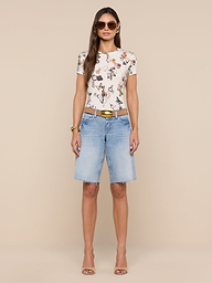 A woman stands facing forward, wearing sunglasses, the bestselling Ressi Fitted Tee featuring a floral graphic, light blue slim-cut knee-length denim shorts, a brown belt, and beige heels against a plain white background.
