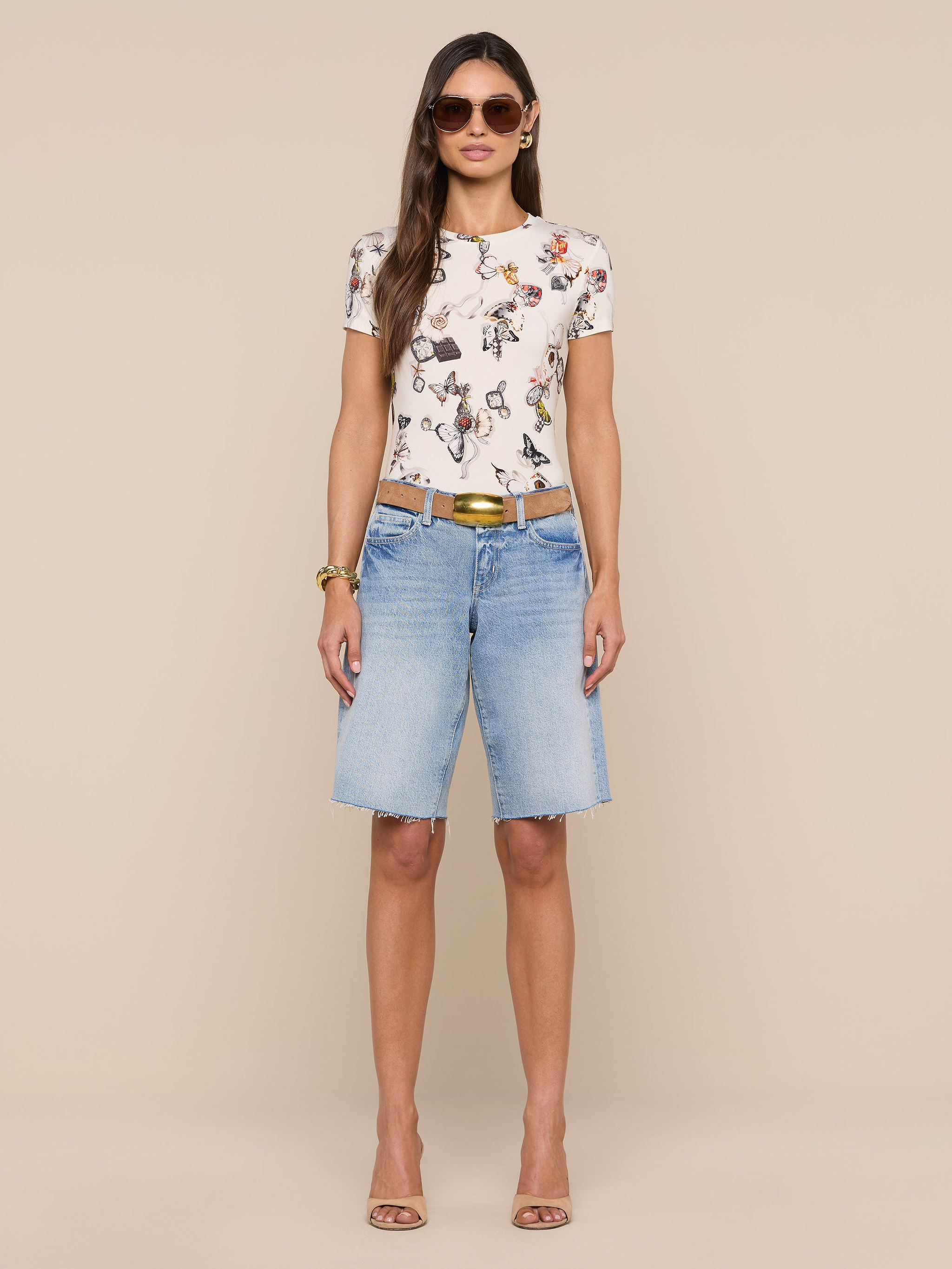 A woman stands facing forward, wearing sunglasses, the bestselling Ressi Fitted Tee featuring a floral graphic, light blue slim-cut knee-length denim shorts, a brown belt, and beige heels against a plain white background.