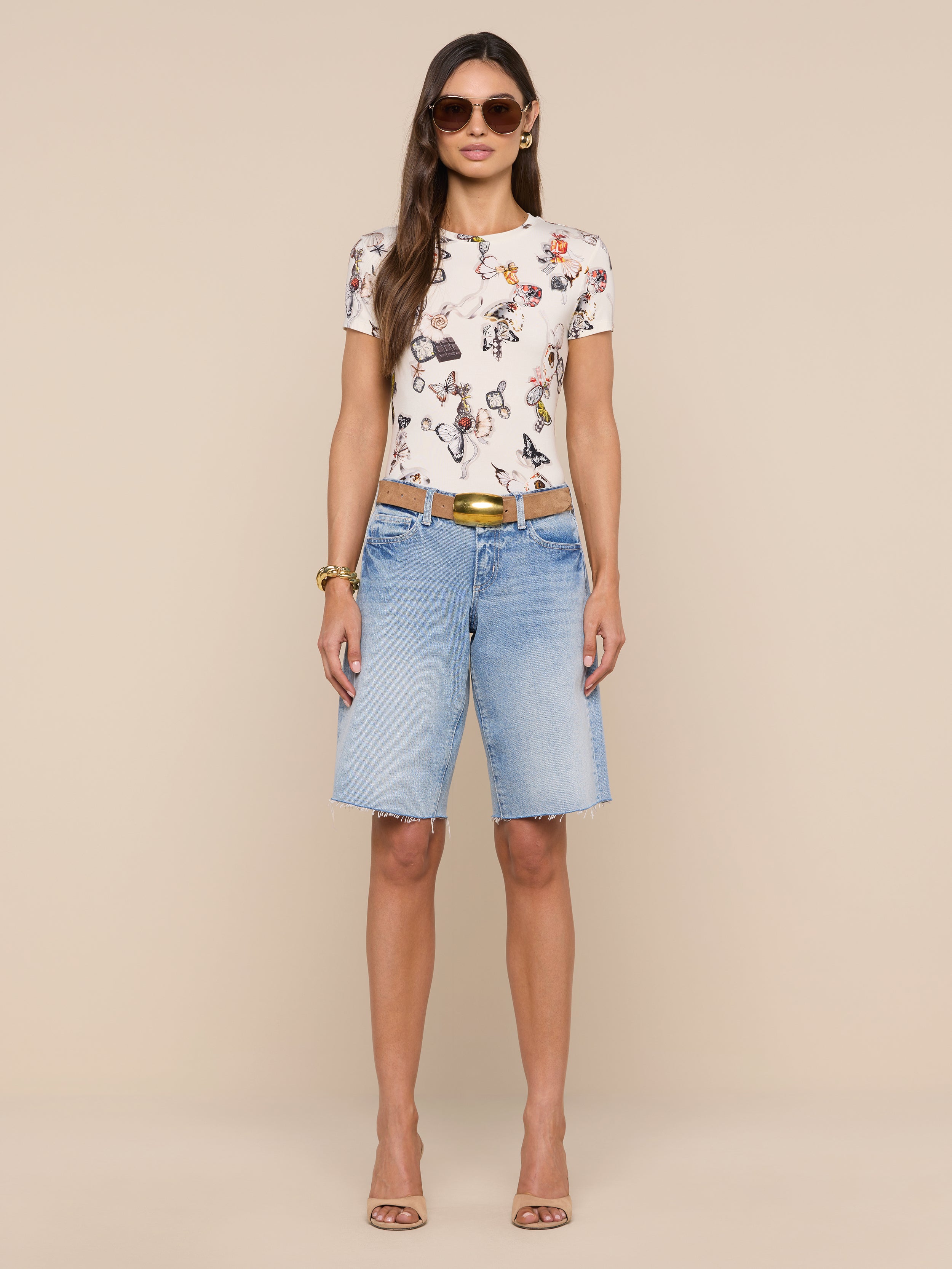 A woman stands facing forward, wearing sunglasses, the bestselling Ressi Fitted Tee featuring a floral graphic, light blue slim-cut knee-length denim shorts, a brown belt, and beige heels against a plain white background.
