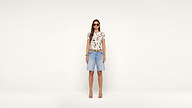 A woman wearing sunglasses, a floral t-shirt, denim knee-length shorts, and heels stands against a plain white background, facing forward and smiling.