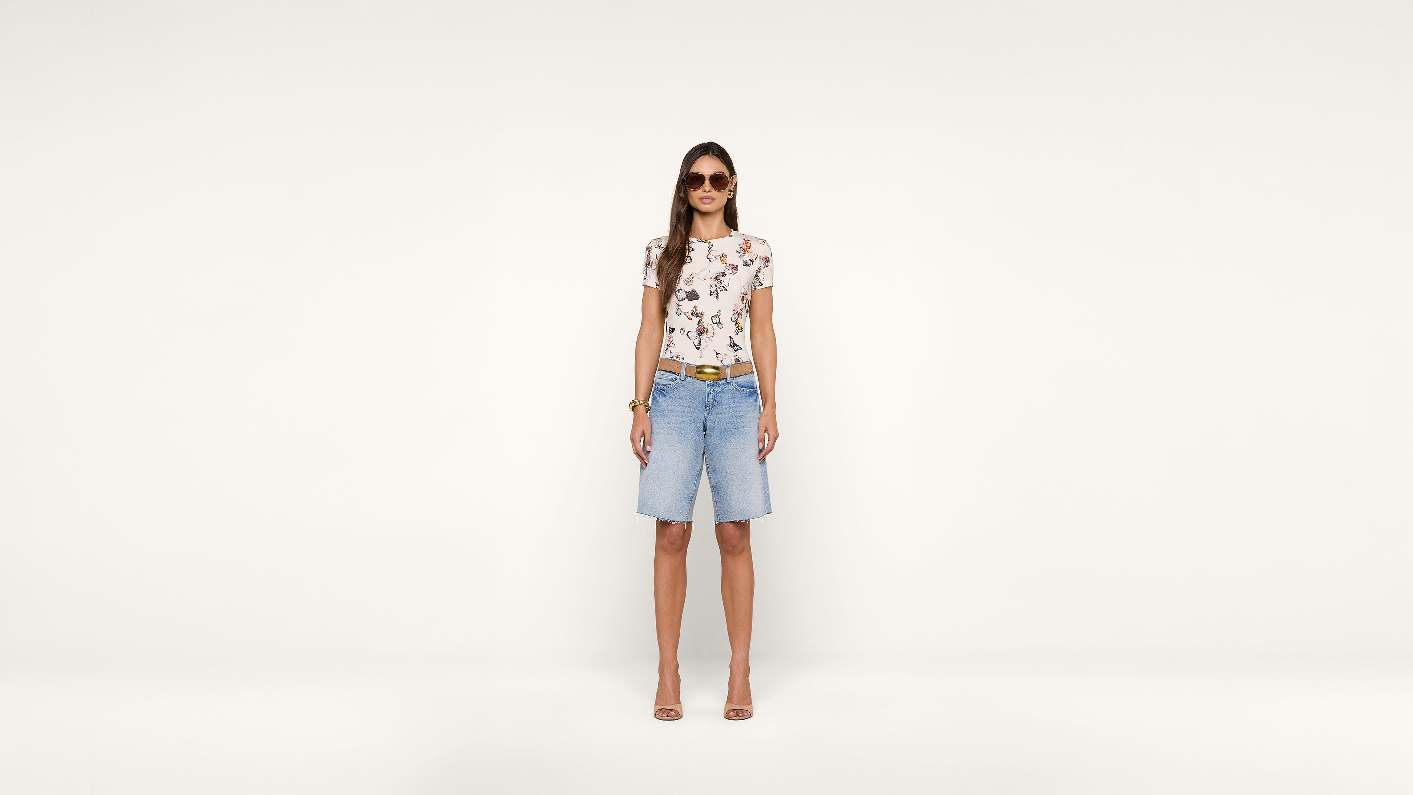 A woman wearing sunglasses, a floral t-shirt, denim knee-length shorts, and heels stands against a plain white background, facing forward and smiling.