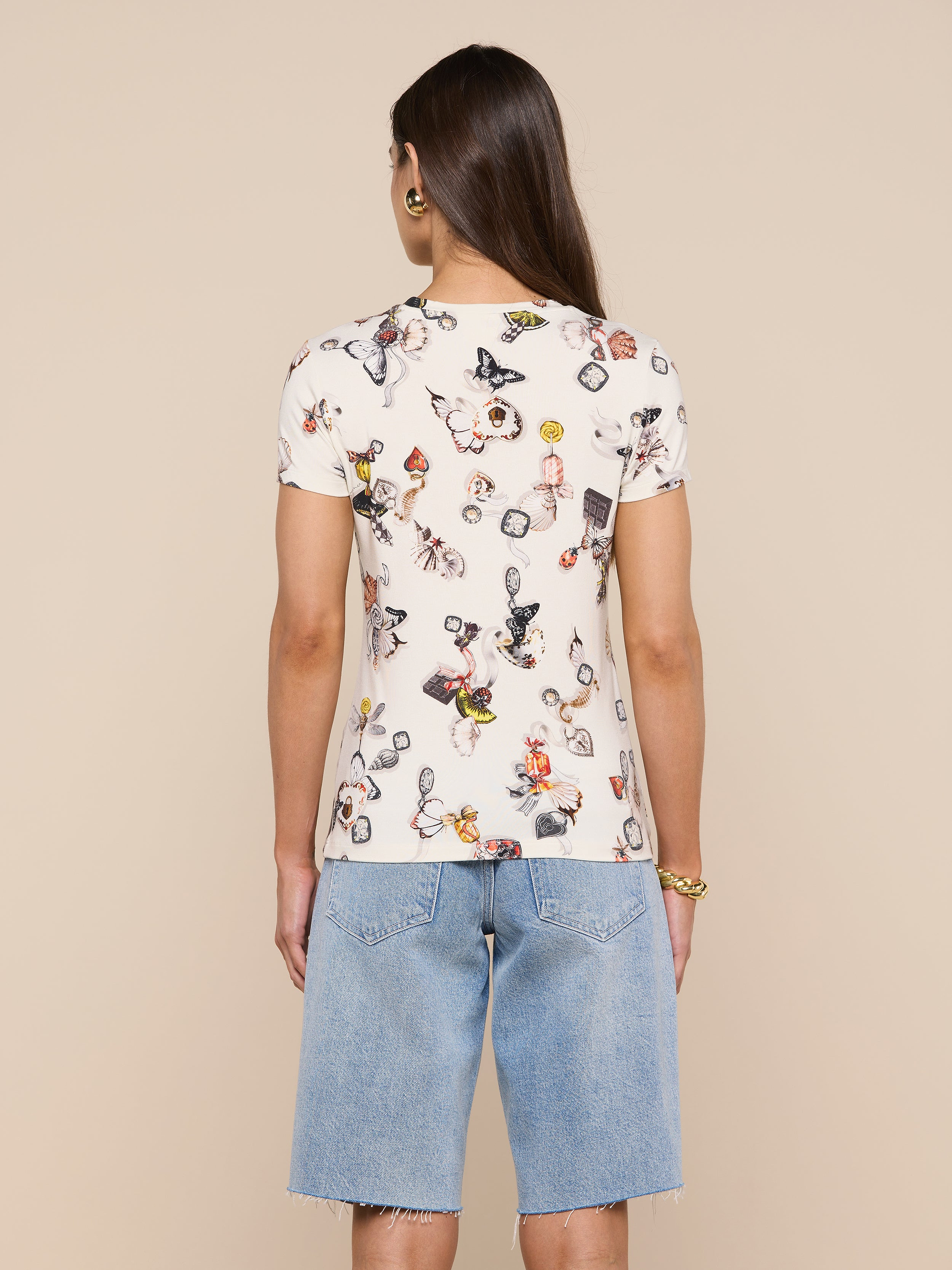 A person with long brown hair wears the bestselling Ressi Fitted Tee in printed white stretch jersey knit and light blue knee-length denim shorts, standing with their back to the camera against a plain light background.