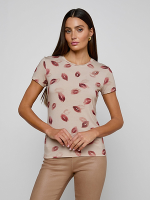 A woman with long brown hair wears the Ressi Fitted Tee—a slim beige t-shirt with a red lipstick kiss print—and beige pants, standing against a light gray background, looking forward with a neutral expression.