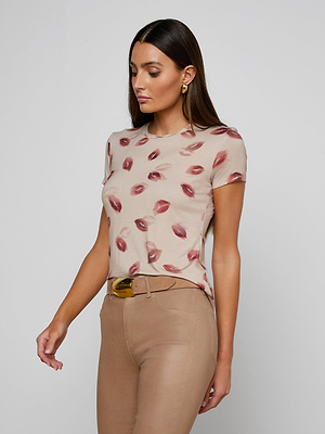 A woman with long brown hair wears the bestselling Ressi Fitted Tee in beige stretch jersey knit with a red lips print, paired with beige pants. She looks down and to the side against a plain light background.