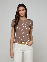 A woman with long brown hair wears the Ressi Fitted Tee, a slim-cut brown short-sleeve shirt with white polka dots, paired with white pants. She stands against a plain light background, one hand in her pocket.
