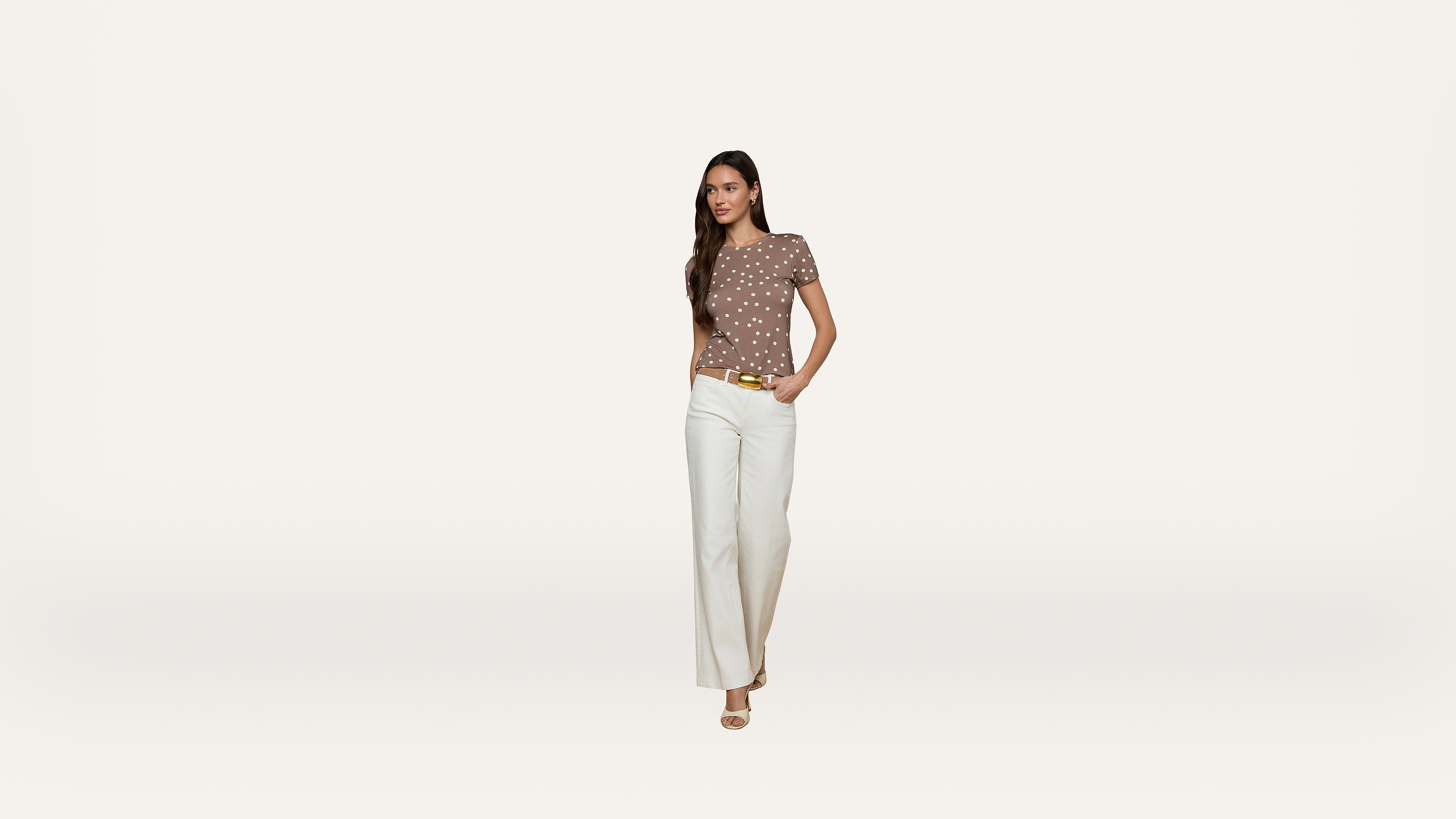 A woman stands against a plain white background, wearing a brown polka dot top, white wide-leg pants, and beige heels, with one hand in her pocket.