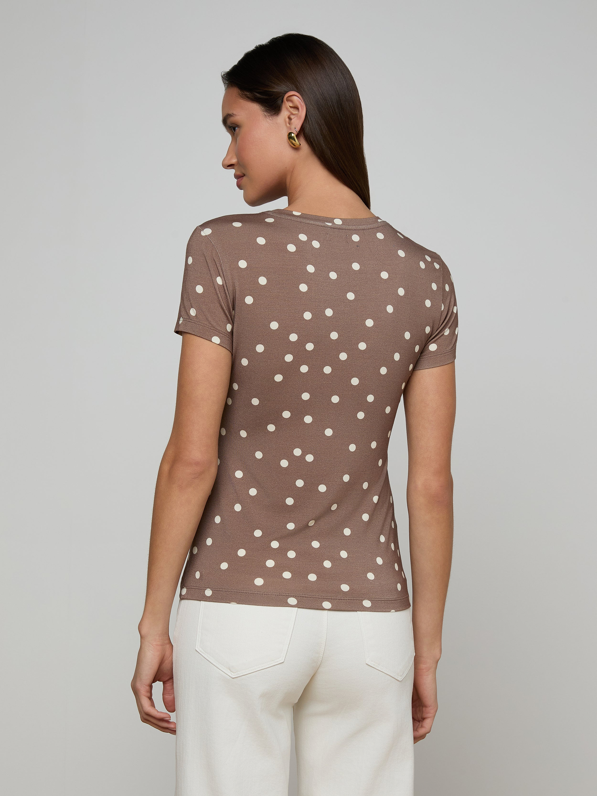 A woman stands facing away, wearing the Ressi Fitted Tee—a slim cut brown short-sleeve shirt with white polka dots—and white pants, set against a plain light background.