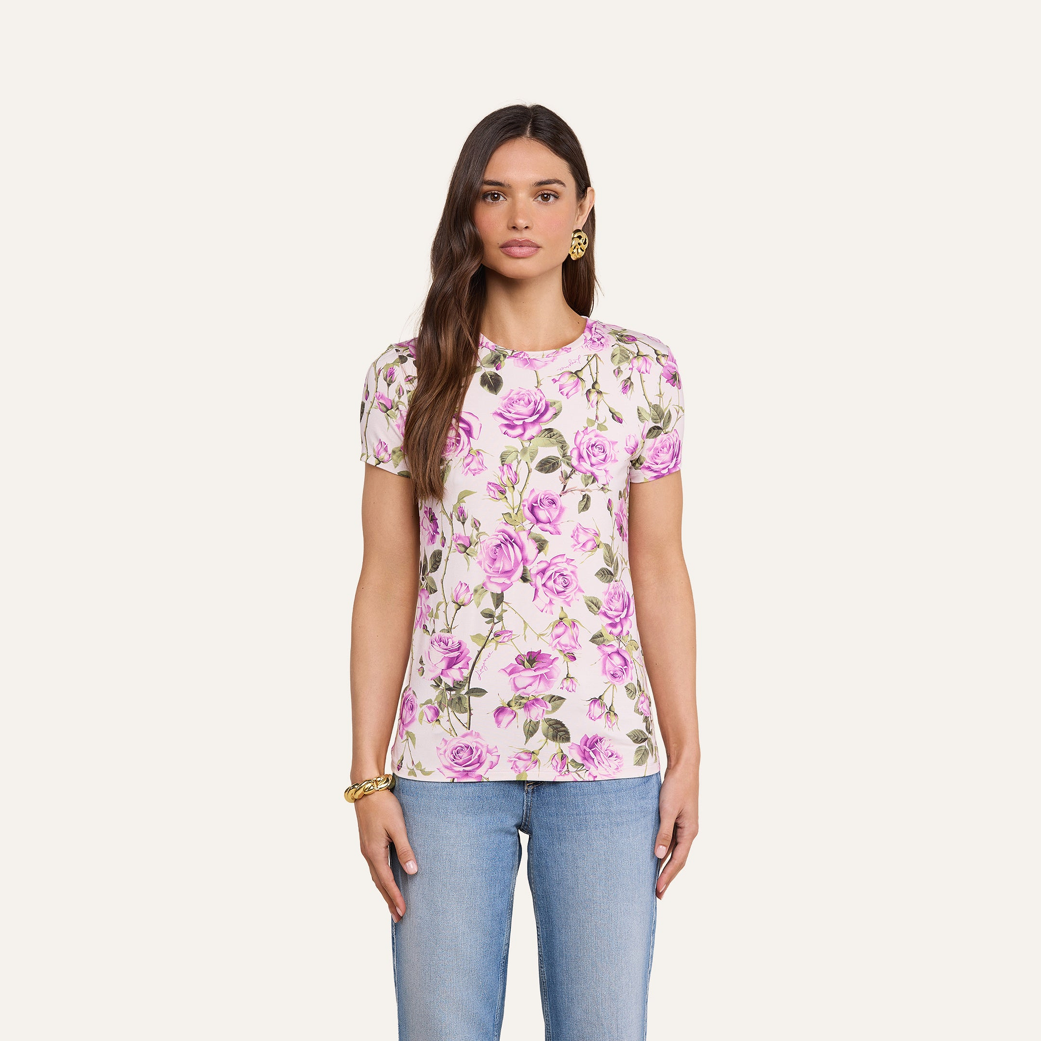 Una donna dai lunghi capelli castani indossa la maglietta Ressi Fitted Tee, best seller in jersey elasticizzato con una stampa di rose rosa e verdi, abbinata a jeans blu e gioielli dorati, in piedi su uno sfondo beige.
