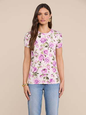 A woman with long brown hair wears the bestselling Ressi Fitted Tee in stretch jersey with a pink and green rose print, paired with blue jeans and gold jewelry, standing against a beige background.
