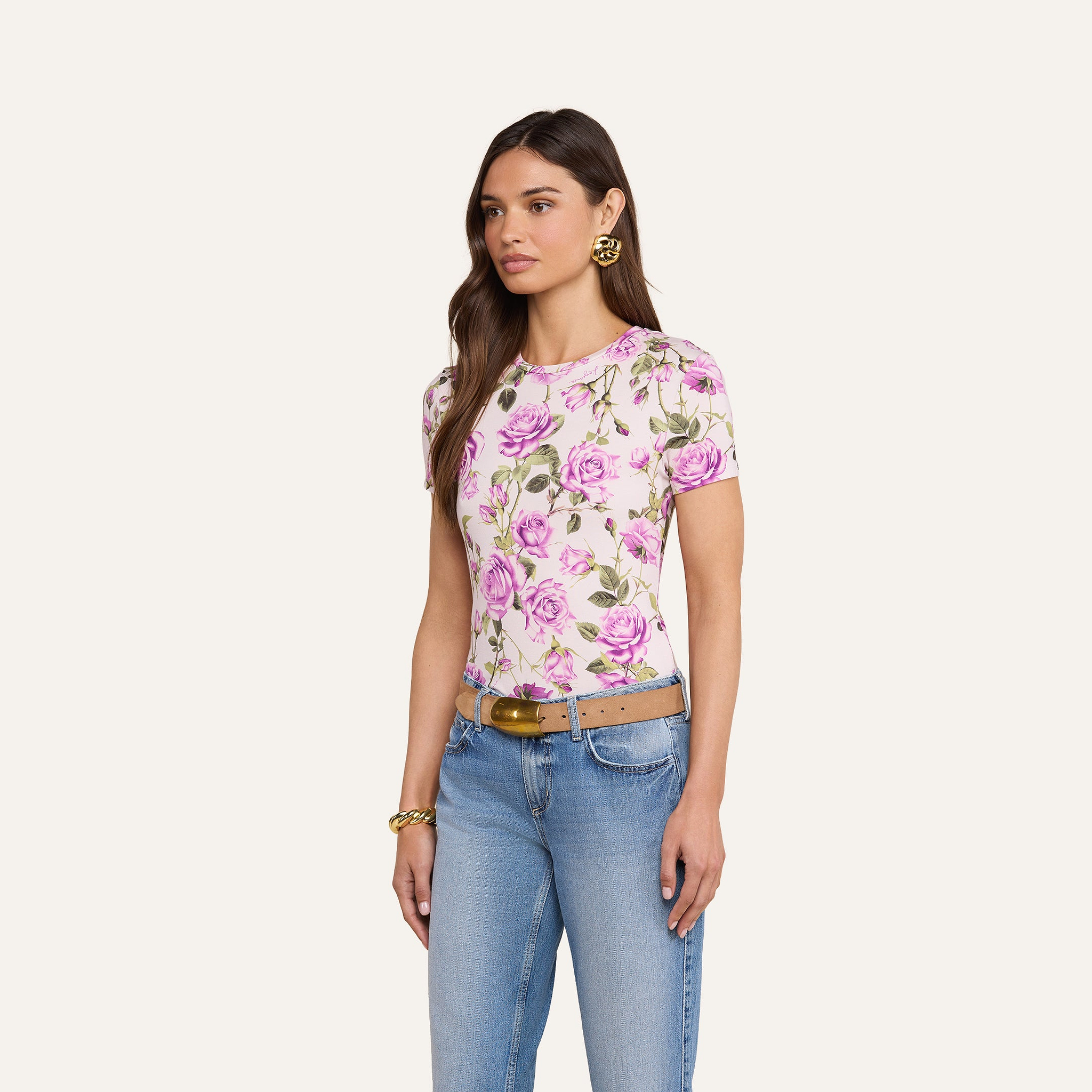Una donna è in posa su uno sfondo beige e indossa la maglietta Ressi Fitted Tee, best seller in jersey elasticizzato con stampa di rose rosa, jeans blu, cintura color cuoio, orecchini dorati e un braccialetto dorato.