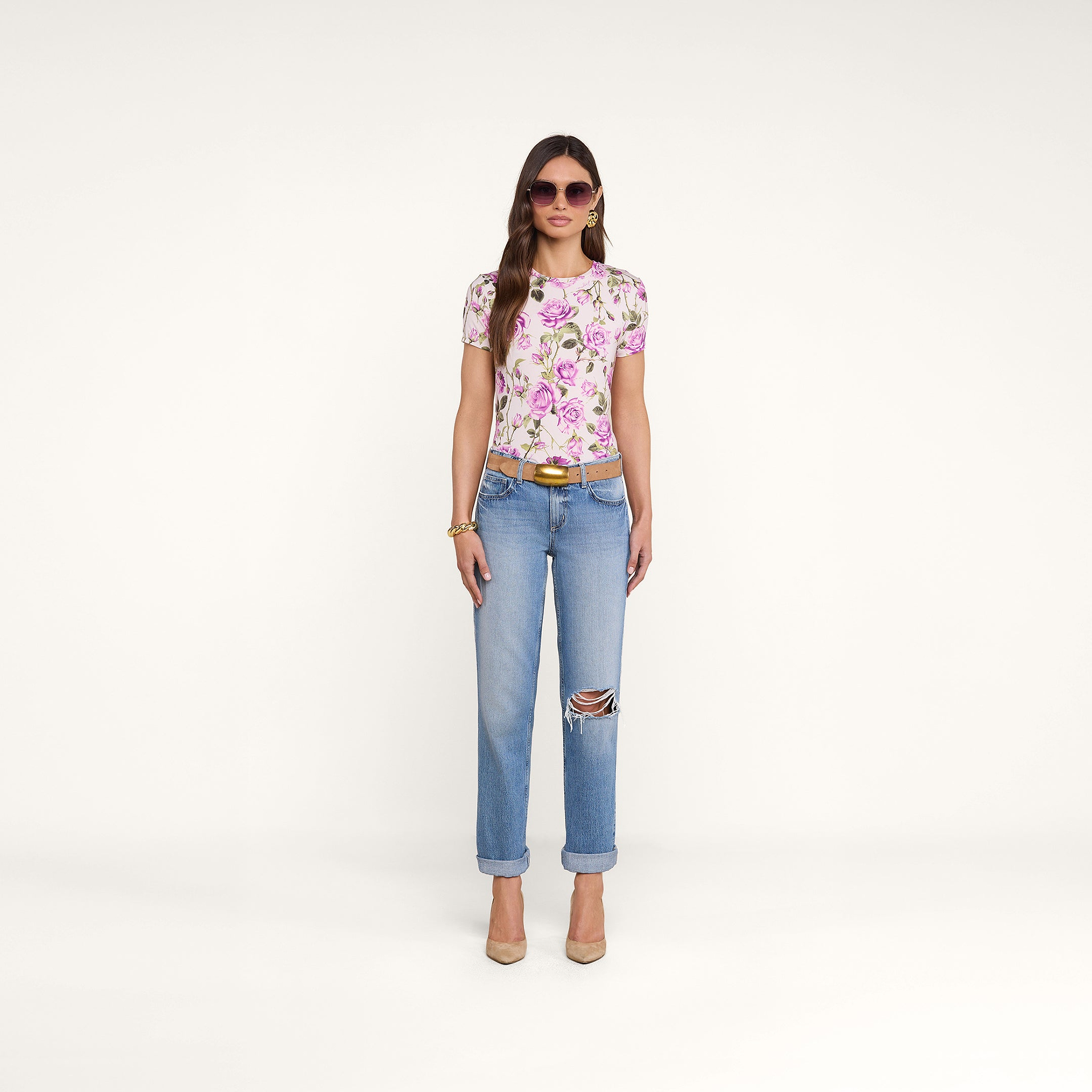 Una donna dai capelli lunghi e con occhiali da sole posa su uno sfondo beige, indossando jeans blu strappati e la maglietta Ressi Fitted Tee, best seller della collezione, realizzata in jersey elasticizzato con stampa di rose bianche e rosa, oltre a gioielli dorati, una cintura marrone e tacchi beige.
