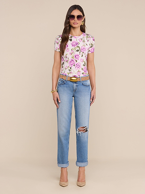 A woman with long hair and sunglasses poses against a beige background, styled in blue ripped jeans and the bestselling Ressi Fitted Tee in a white and pink rose print stretch jersey, plus gold jewelry, a brown belt, and beige heels.