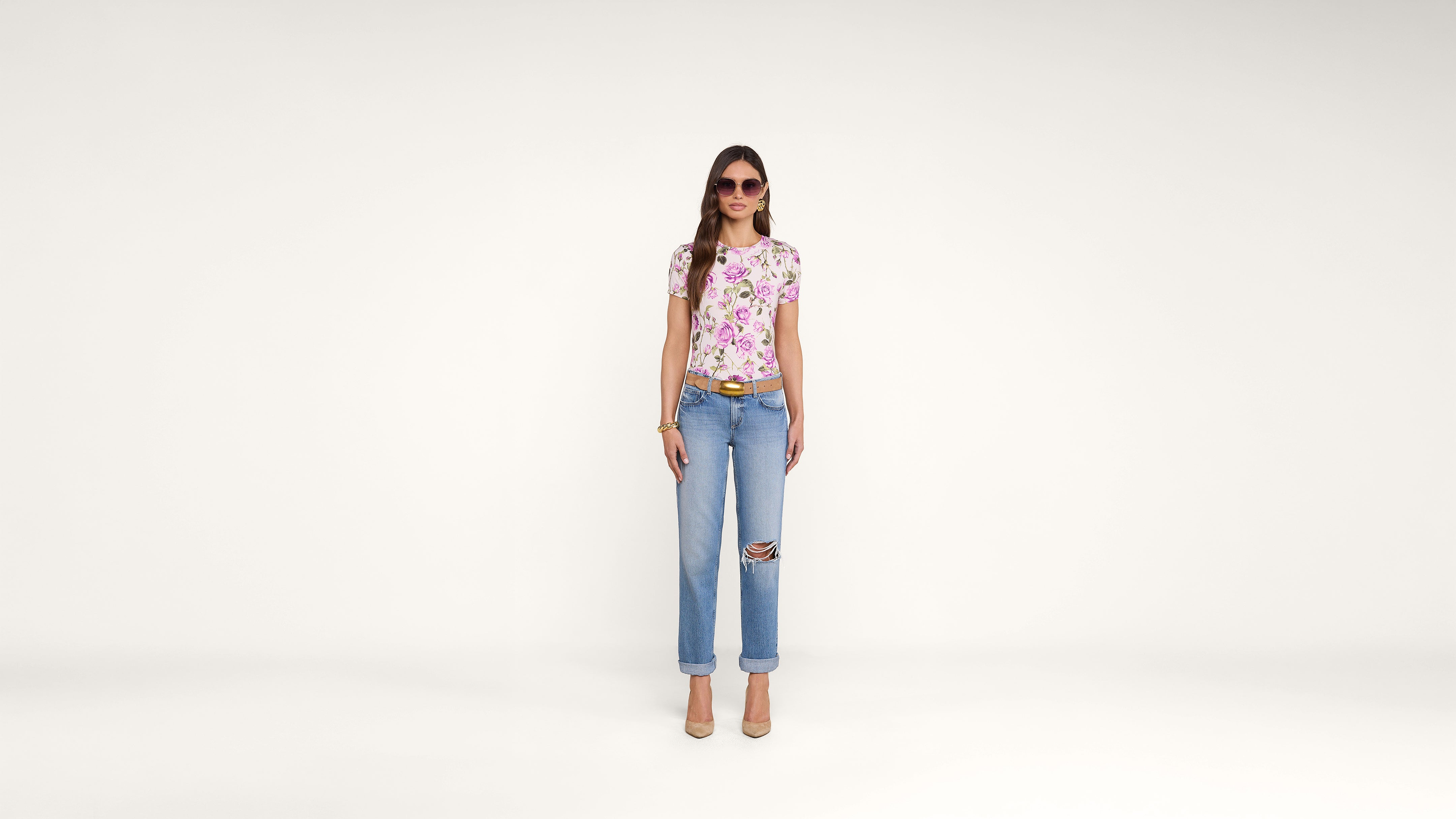 A woman wearing sunglasses, a floral t-shirt, ripped blue jeans, and beige heels stands against a plain white background.