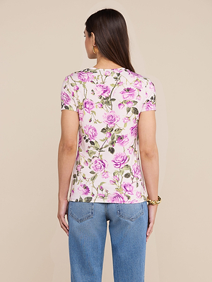 A woman with long brown hair, wearing the Ressi Fitted Tee in stretch jersey knit with a pink and green floral rose print, paired with blue jeans, faces away from the camera against a plain beige background.