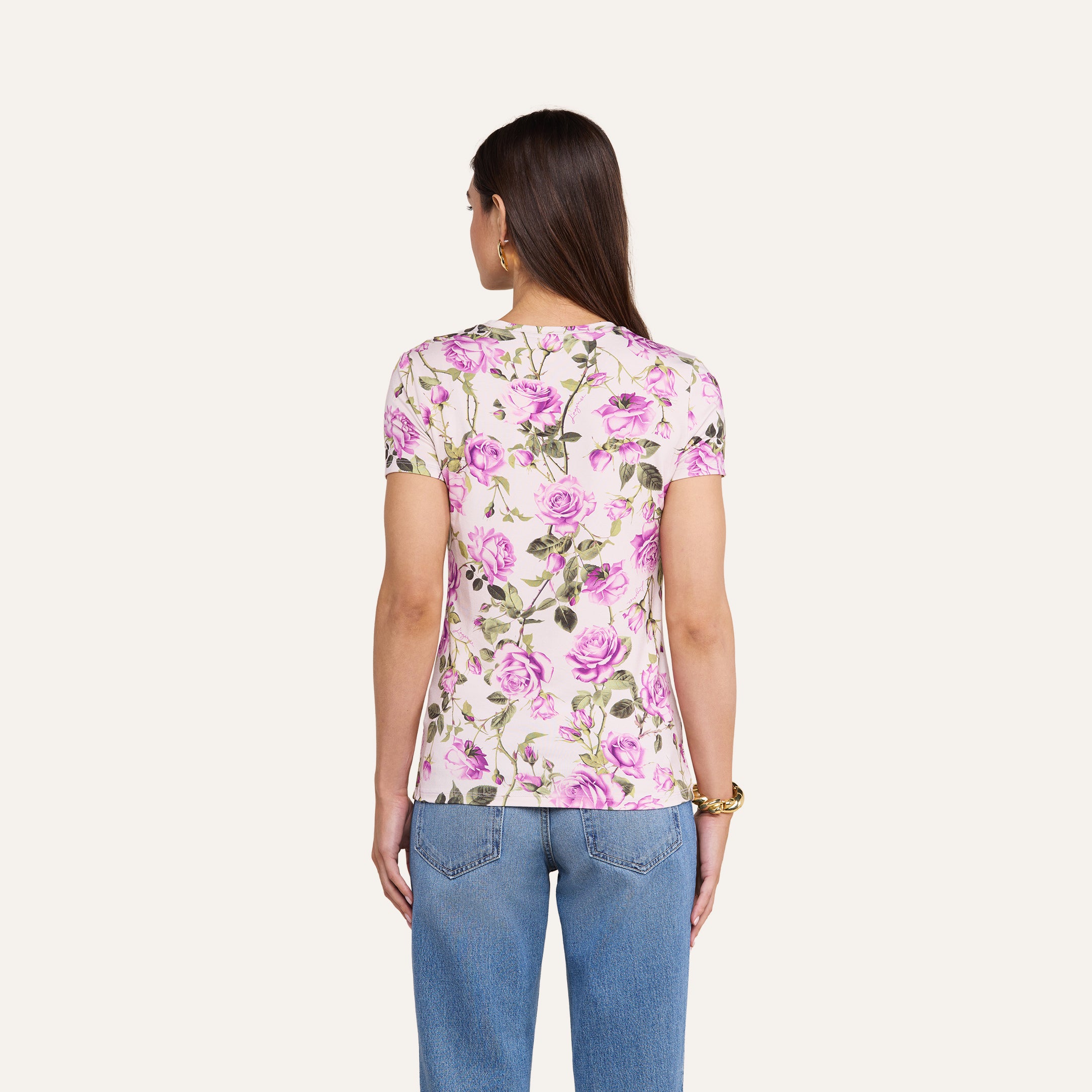 Una donna dai lunghi capelli castani, che indossa la maglietta aderente Ressi in jersey elasticizzato con una stampa floreale di rose rosa e verdi, abbinata a jeans blu, è di spalle alla fotocamera su uno sfondo beige a tinta unita.
