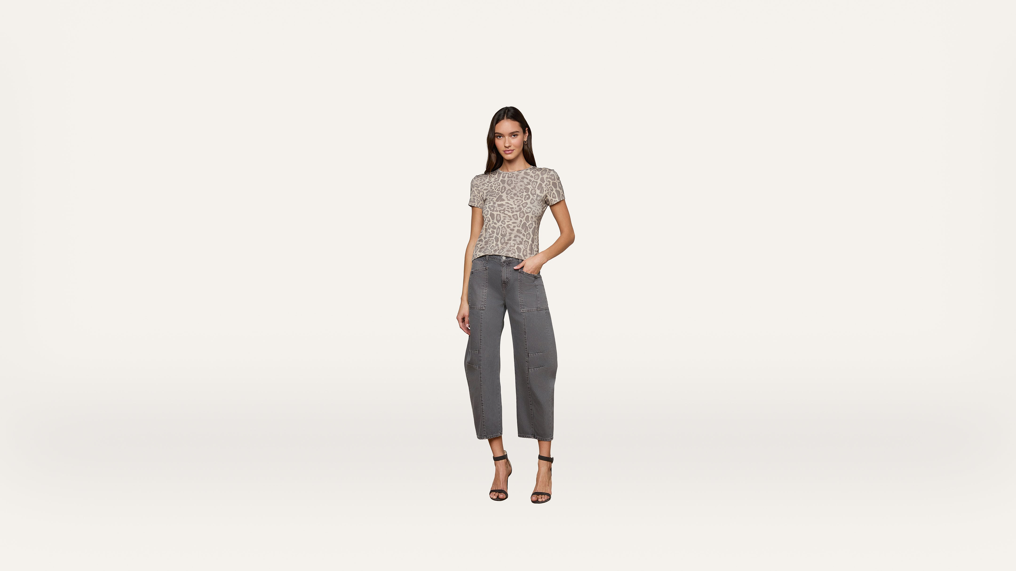 A woman stands against a plain white background, wearing a short-sleeve patterned top, gray cropped jeans, and black high-heeled sandals. She has one hand in her pocket and is looking at the camera.