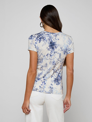 A woman with long brown hair and hoop earrings stands with her back to the camera against a light background, wearing the bestselling Ressi Fitted Tee in a blue and white floral paisley print, paired with white pants.