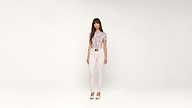 A woman with long brown hair stands against a plain white background, wearing a patterned short-sleeve shirt, white pants, and open-toed heels.