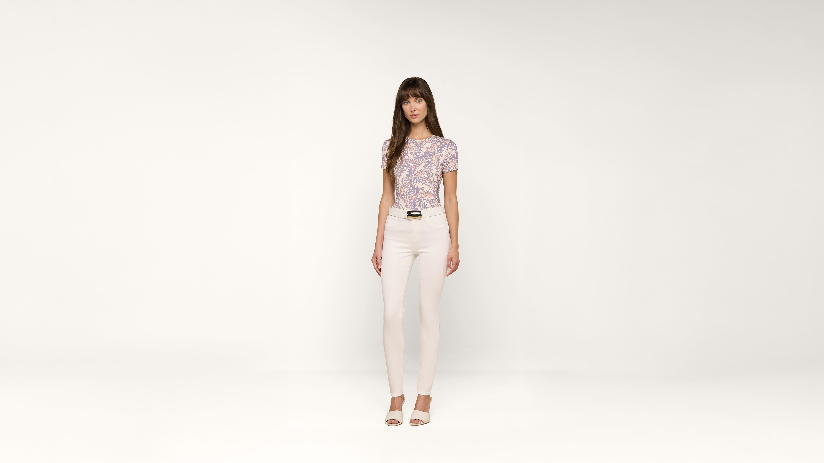 A woman with long brown hair stands against a plain white background, wearing a patterned short-sleeve shirt, white pants, and open-toed heels.