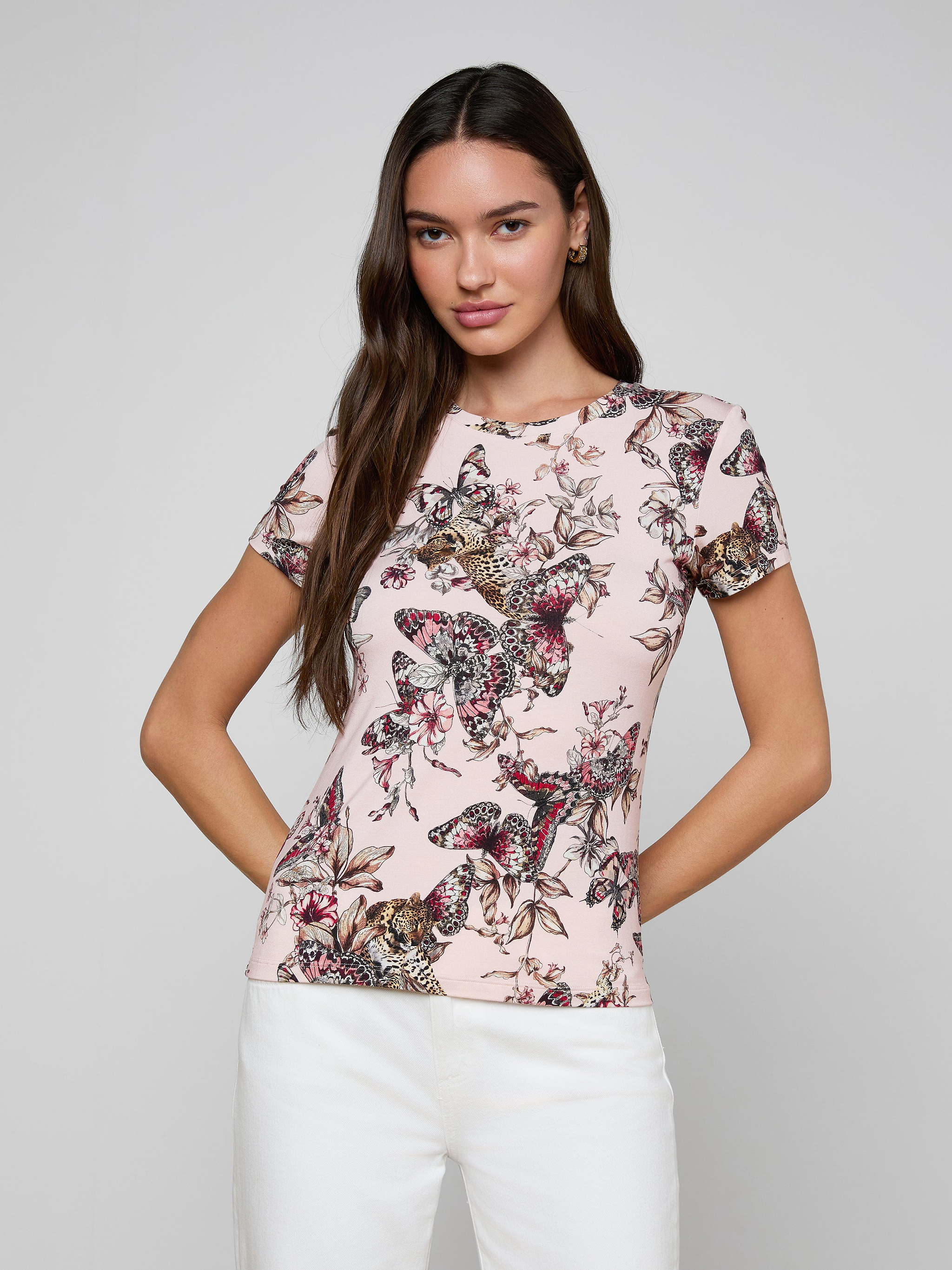 A woman stands against a plain background wearing the bestselling Ressi Fitted Tee, a light pink short-sleeve top crafted from stretch jersey knit and adorned with a vibrant butterfly jungle print, paired with white pants.