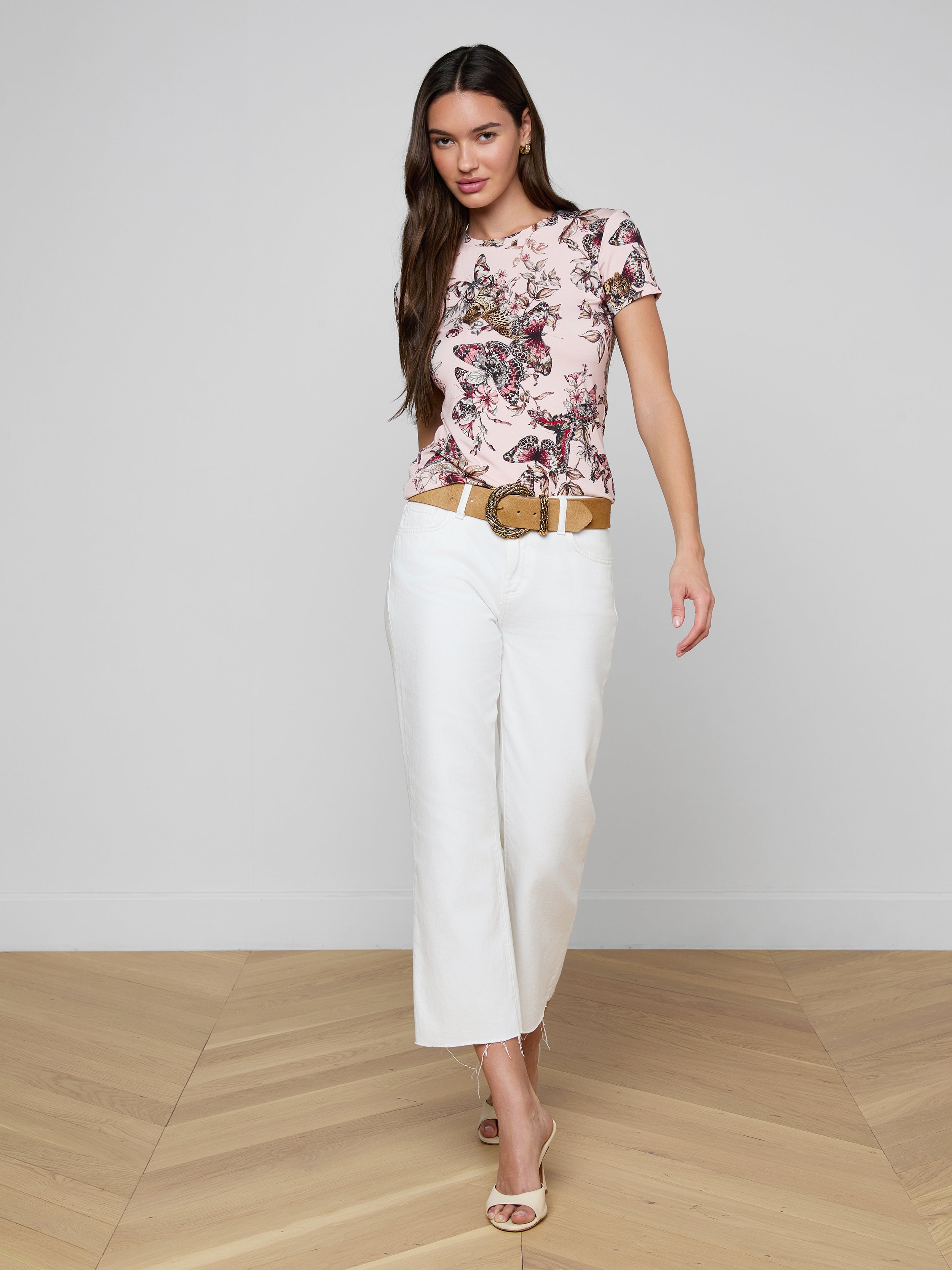 Woman wearing the bestselling Ressi Fitted Tee with a butterfly jungle print, white wide-leg pants, beige belt, and open-toe heels stands against a plain, light background.