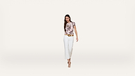 A woman with long hair wearing a floral top, white pants, a tan belt, and heeled sandals is walking forward against a plain white background.