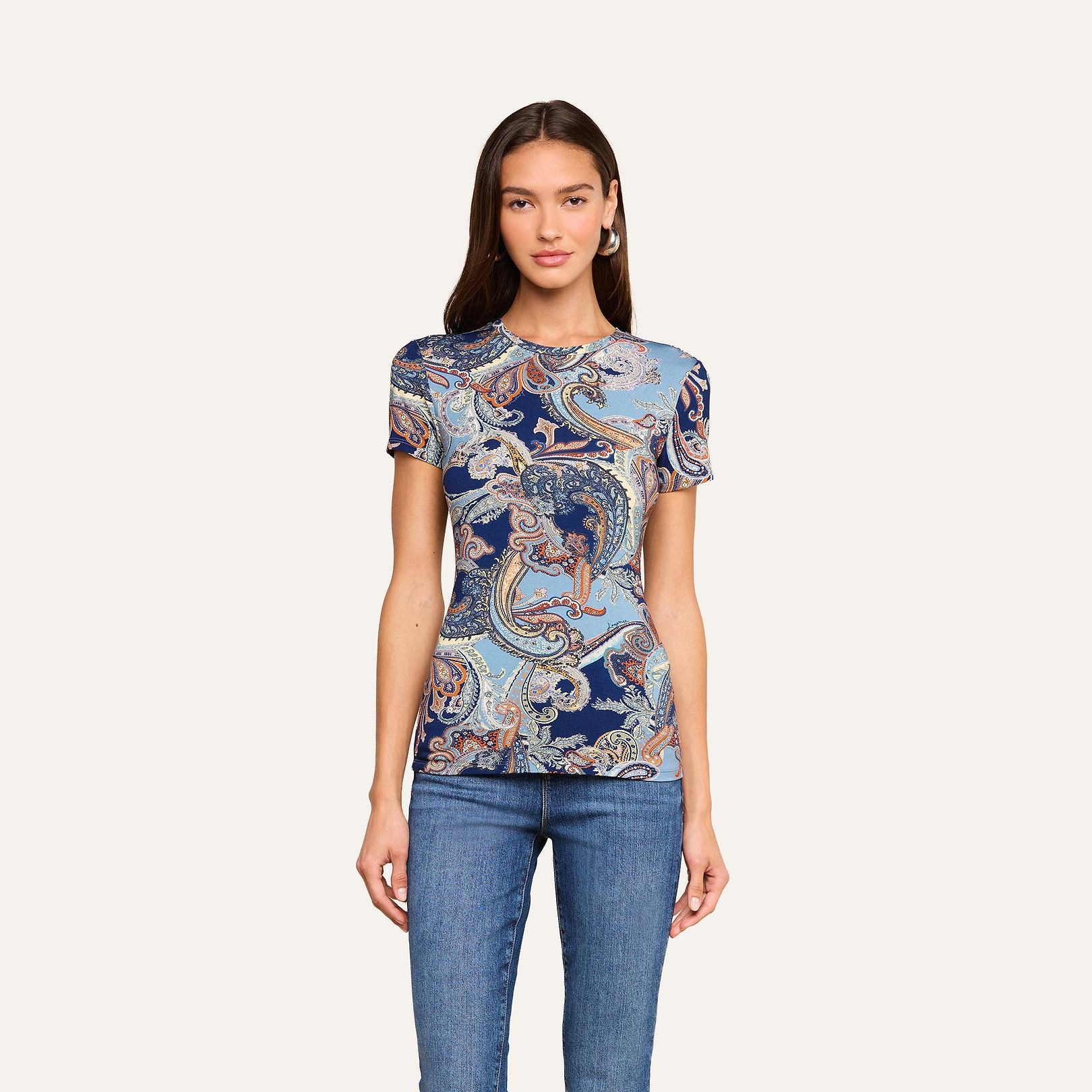 Indossa la maglietta aderente Ressi — un top a maniche corte con motivo paisley blu notte — e, con indosso jeans blu dal taglio slim, una donna è ritratta di fronte, su uno sfondo beige, con un’espressione neutra e le mani lungo i fianchi.