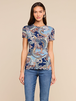 Wearing the Ressi Fitted Tee—a short sleeve top with a midnight blue paisley pattern—a woman in slim fit blue jeans stands facing forward against a beige background, maintaining a neutral expression with her hands at her sides.