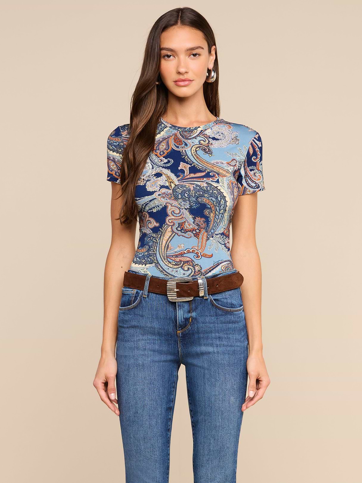 Donna dai lunghi capelli castani che indossa la maglietta aderente Ressi con motivo paisley blu notte, abbinata a jeans blu e una cintura marrone, in piedi su uno sfondo beige a tinta unita.