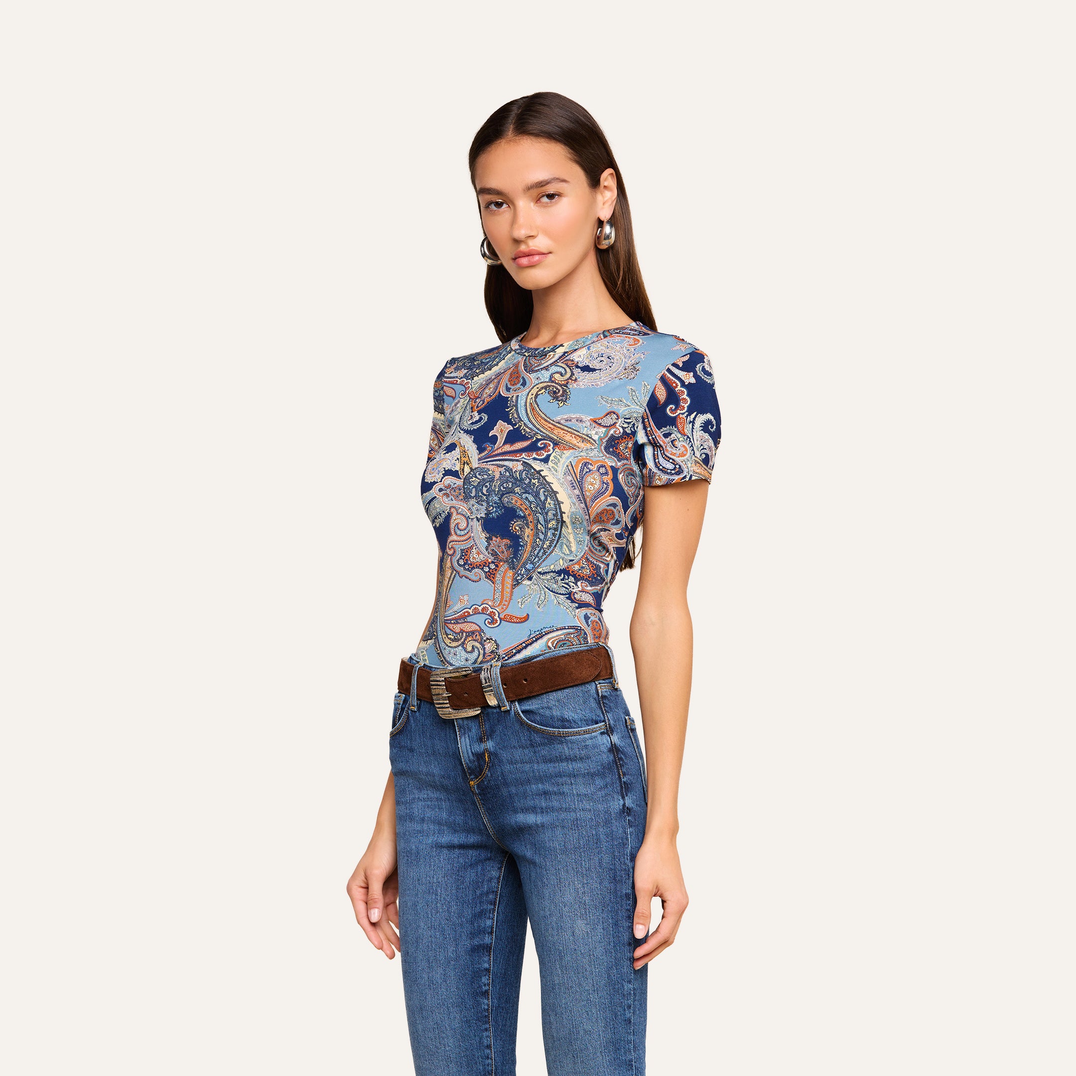 Une femme portant un t-shirt ajusté Ressi bleu nuit à motif cachemire, un jean bleu et une ceinture marron, debout devant un fond beige uni. Elle a de longs cheveux bruns et porte des boucles doreilles créoles argentées.